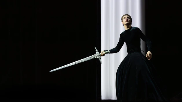 Image of a scene from “Romeo + Juliet” by Marcos Morau with the Opera Ballet Vlaanderen, showing a dancer dressed in black standing in front of a white curtain, holding out a silver sword. 
