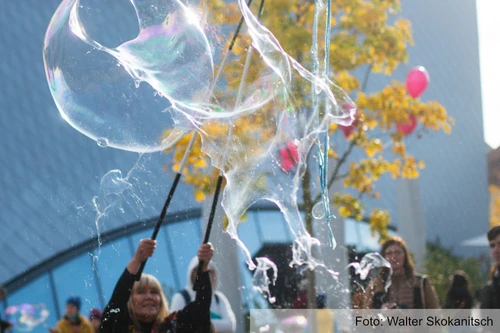 Eine Person lässt im Freien große Seifenblasen entstehen; im Hintergrund stehen Menschen, herbstlich gefärbte Bäume und einige rote Luftballons.