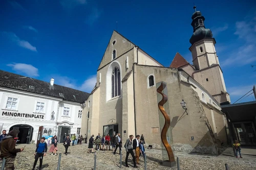 Historischer Platz mit gotischer Kirche, moderner Skulptur und Fußgängern bei sonnigem Wetter.