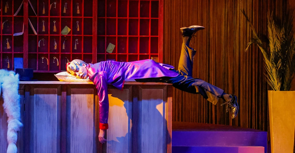 Mann liegt auf Hotelbar und schläft auf einen Polster, hat ein Bein nach oben abgewinkelt, links hängt von der Bar ein Pelzschal, im Hintergrund Schlüsselfächer