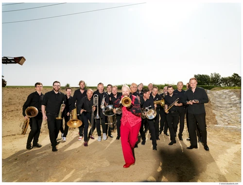 Eine große Gruppe Musiker mit Blasinstrumenten, vorne ein Mann in roter Hose und rotem  Gilet mit Flügelhorn