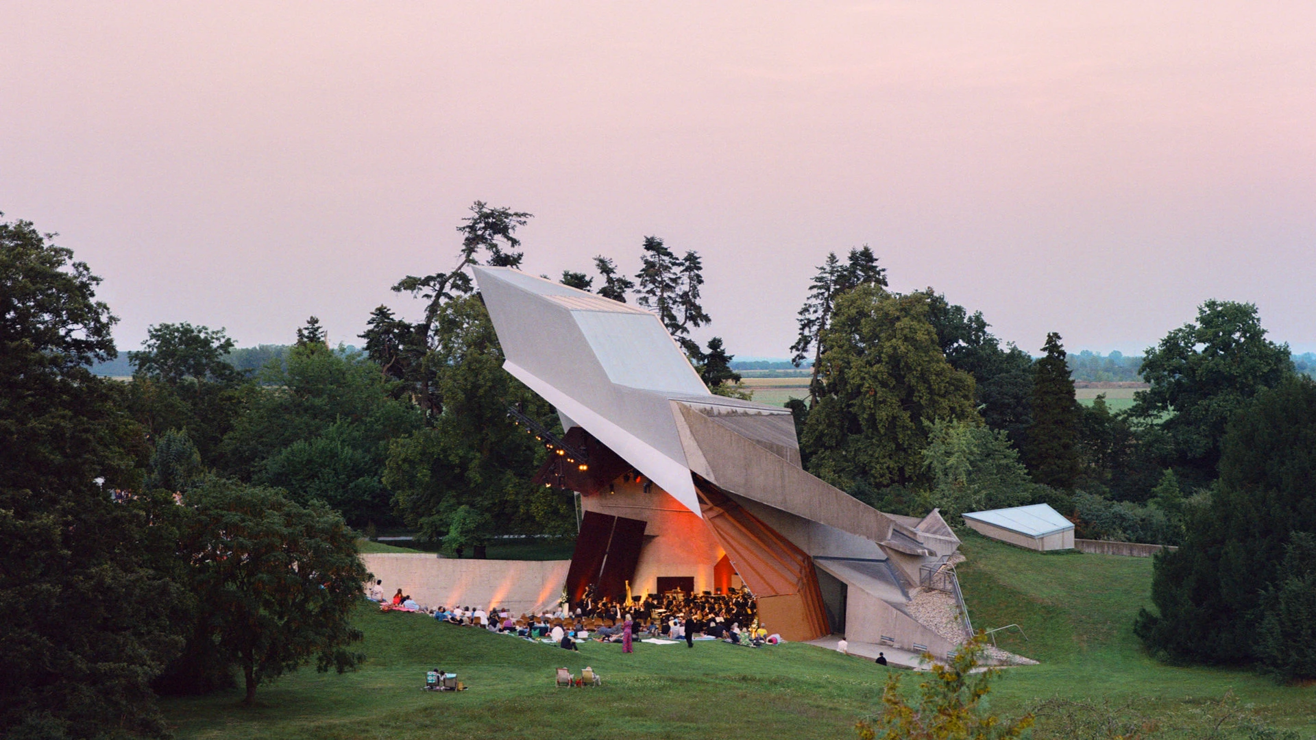 Architektonisch außergewöhnliche Freiluftbühne mit Konzertbesuchern im Grünen bei Sonnenuntergang.
