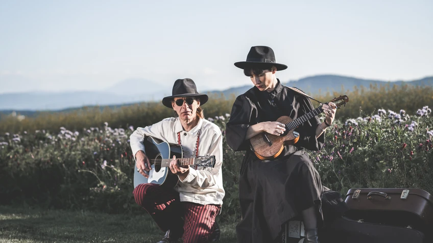 Ernst Molden und Sigrid Horn sitzen mit breitrandigen Hüten in einer blühenden Wiesenlandschaft vor Bergkulisse und spielen Gitarre bzw. Ukulele, während neben ihnen offene Instrumentenkoffer liegen.
