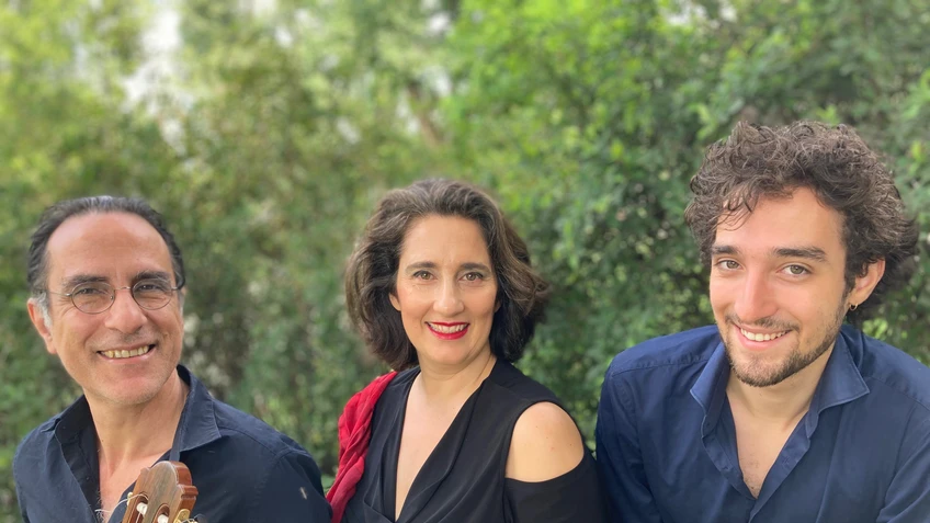 Gruppenfoto Karmon Vounelakos Trio. Drei Personen stehen im Freien mit Bäumen im Hintergrund und lächeln in die Kamera. In der Mitte steht Shira Karmon.