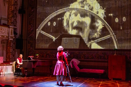 A person in a red stage costume stands in the centre of a theatre stage in front of a large-scale projection. The stage set is bathed in warm red light.