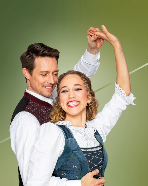Grüner Hintergrund, davor Mann und Frau in Tanzpose, beide tragen Tracht. Der Mann blickt Frau lächelnd an, Frau lacht in die Kamera.