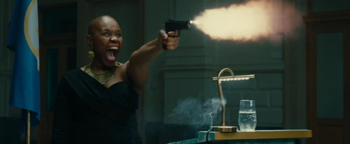 A still from the short film ‘Two to Tango’ by Dimitri Sterkens. It shows a woman in a black dress in a professional setting (with a backdrop and a lectern next to or behind her) firing a gun, her mouth wide open in a scream.