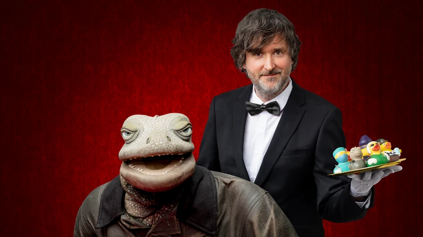 Michael Hatzius steht im Smoking vor einem roten Hintergrund und hält ein Tablett mit bunten Plastikenten, während neben ihm die Echse in brauner Kleidung sitzt und eine Zigarre in der Hand hält.