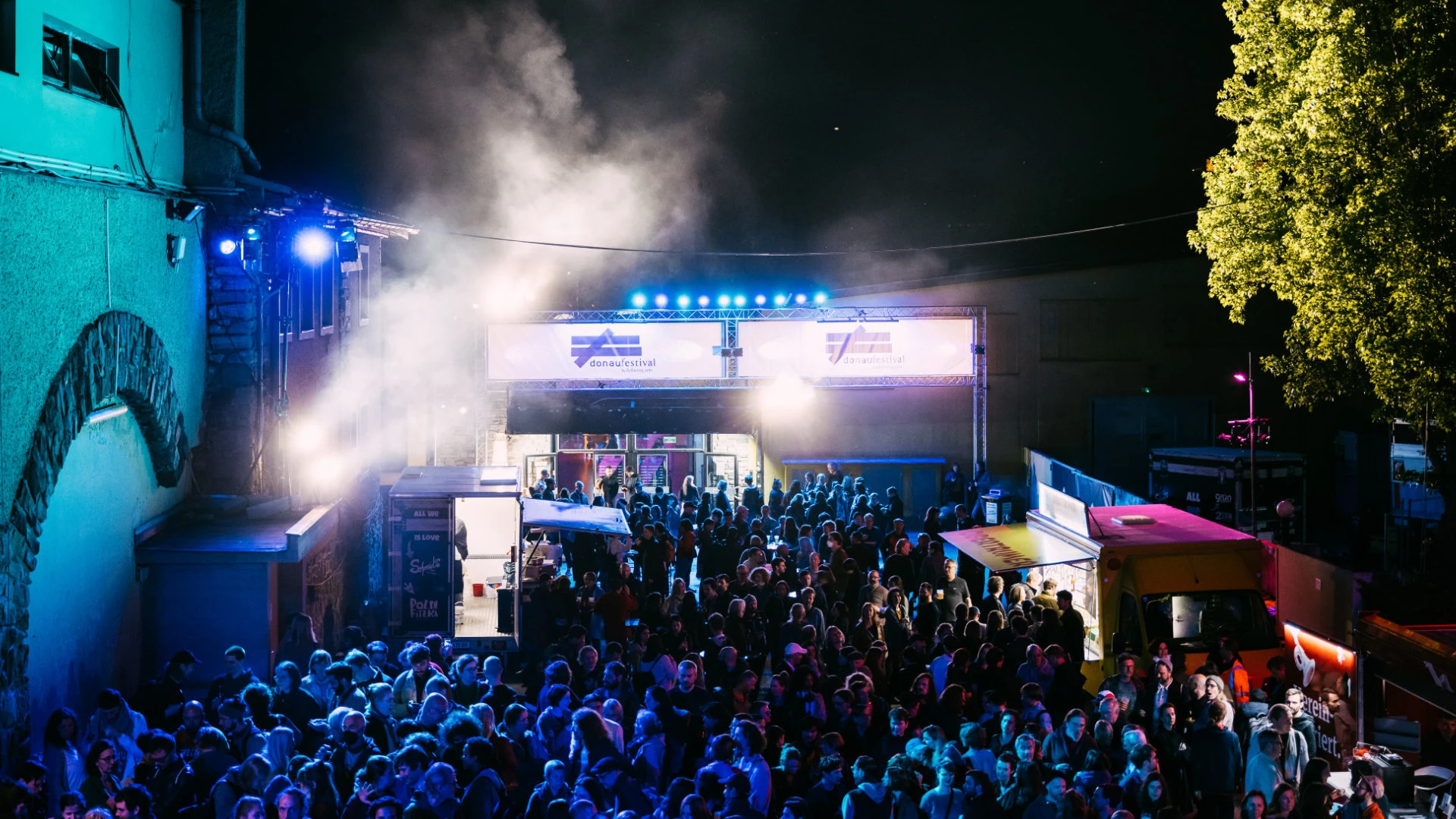 Menschenmenge vor dem Eingang eines nächtlichen Musikfestivals mit Lichtinstallation, Foodtrucks und Bühnenbeleuchtung.