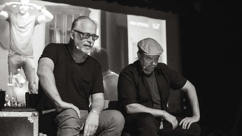 Peter Hörmanseder und Robert Stachel sitzen auf einer dunklen Bühne mit amüsiertem und ernstem Ausdruck nebeneinander, während hinter ihnen auf einer großen Leinwand eine Szene aus einer Nachrichtensendung projiziert wird.