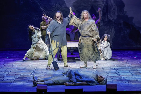 Two men in shabby clothes give each other a high five with their eyes facing forward, the man on the left is holding a shovel, in the background there are some people dancing