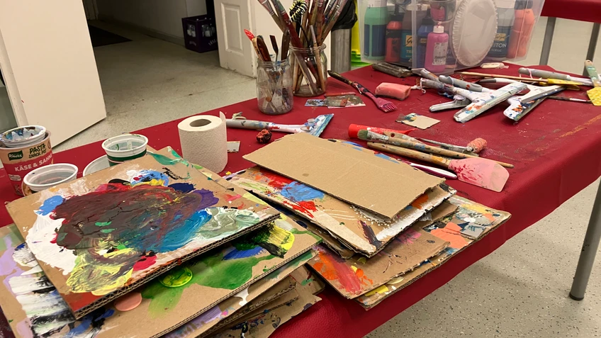 Kreativer Arbeitstisch mit bemalten Kartonplatten, Pinseln, Farben und Malutensilien in einem Atelier- oder Workshopraum.