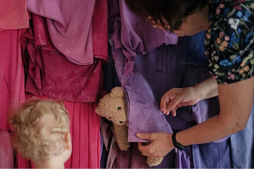 Kind schaut auf Teddybär, der mithilfe von Oma, hinter dem bunten Stoff der Regenbogenwand im KinderKunstLabor hervorlugt.