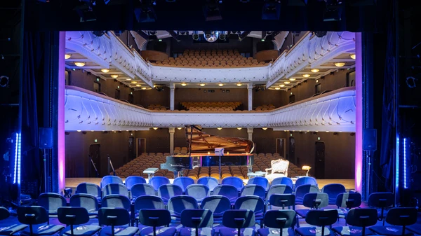 Leere Stuhlreihen auf einer Bühne mit Blick in einen beleuchteten Theatersaal, davor ein Flügel