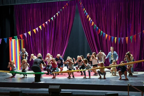 Eine Gruppe Kinder in der Hocke zur Kamera gedreht vor einem violetten Vorhang und bunten Zirkusgirlanden.  