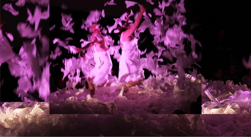 A still from ‘Sous la Neige / Under the Snow’ by Compagnie Les Bestioles. The image shows two performers dressed in white amidst feathers floating through the air against a black stage backdrop.