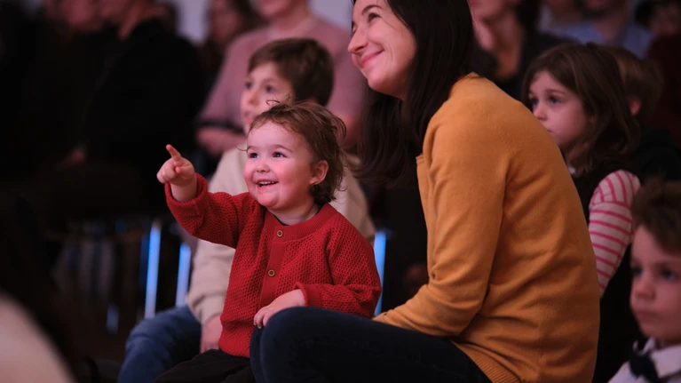 Publikumsfoto vom "Tonspiele"-Projekt "Schnee" - einem Projekt des Musikvermittlungsprogramm des Tonkünstler-Orchester Niederösterreich. Zu sehen sind fröhliche Kinder, die gespannt zuhören und -schauen. Ein Kind sitzt auf dem Schoß seiner Mutter und zeigt in Richtung der Bühne.
