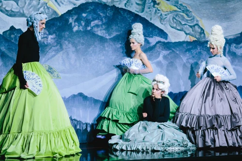 Vier Darstellerinnen in barock inspirierten Kostümen mit Perücken und Fächern auf einer Theaterbühne.