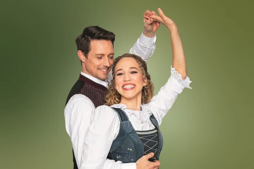 Grüner Hintergrund, davor Mann und Frau in Tanzpose, beide tragen Tracht. Der Mann blickt Frau lächelnd an, Frau lacht in die Kamera.