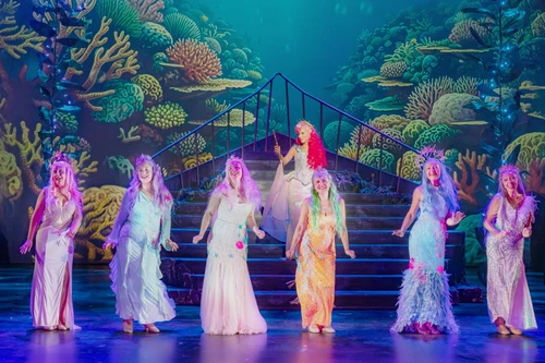 A colourful stage scene features several performers in bright, glittering costumes against a backdrop of coral reefs and marine plants. The group is spread out across the steps of a central staircase. At the centre stands a person wearing a bright pink wig on a raised landing. Blue and turquoise lighting emphasises the underwater atmosphere of the production.