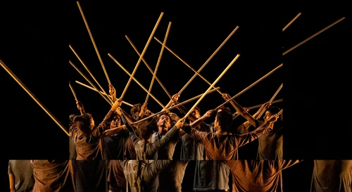 The performers from Collectif Travers & Le Quator Debussy stand close together on a dark stage, all holding sticks aloft with both hands.