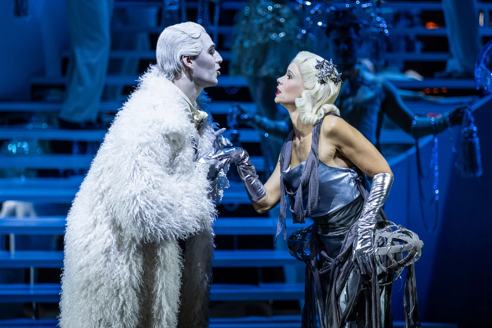 Two stage characters stand facing each other on an illuminated stage, surrounded by a large staircase structure in the background. One figure wears a long, light-coloured, fur-like costume, the other a shiny metallic draped outfit with long gloves and textured fabric elements. Their body language suggests that they are engaged in discussion. The scene is bathed in cool blue light and appears futuristic and festively staged.
