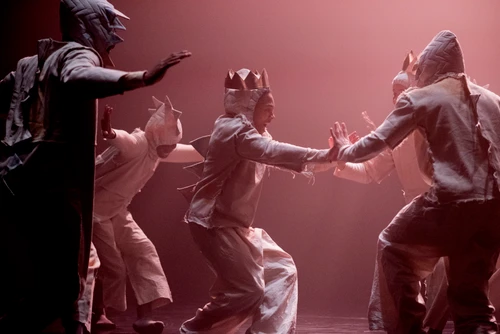 Tänzer:innen der Compagnie Far From The Norm performen "BLKDOG" von Choreograf Botis Seva. Sie tragen weiße Kostüme mit Kapuze, an die Stacheln angenäht sind und tanzen auf einer spärlich in rot beleuchteten Bühne. 