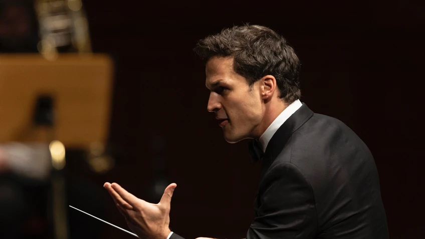 Ein Dirigent mit Dirigierstock im Profil blickt Richtung Orchester, die Hand  ausdrucksvoll angehoben