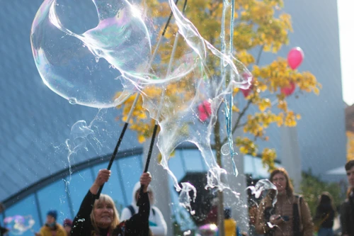 Eine Person lässt im Freien große Seifenblasen entstehen; im Hintergrund stehen Menschen, herbstlich gefärbte Bäume und einige rote Luftballons.