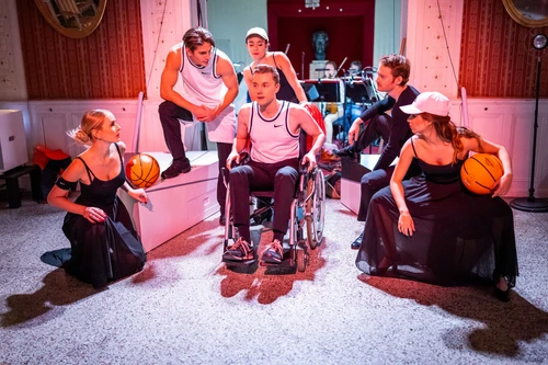 Eine Gruppe junger Menschen, teils mit Basketbällen, teils in Sportkleidung, sitzt rings um einen jungen Mann, der im Rollstuhl sitzt; alle Blicke sind auf die zentrale Figur gerichtet