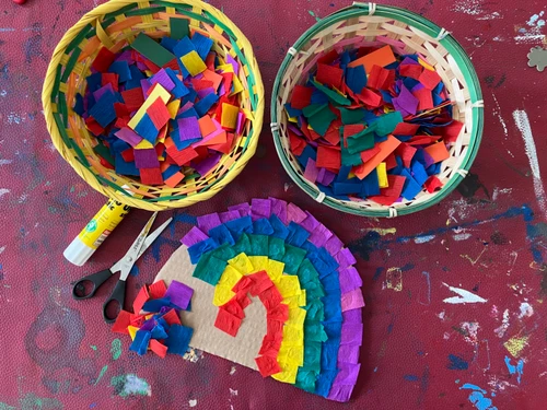 Zwei Körbe mit bunten Papierstücken, daneben eine Schere, ein Klebestift und eine Regenbogen‑Collage auf bemaltem Tisch.