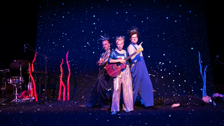 Drei als fantasievolle Weltraumwesen verkleidete Musikerinnen und Musiker stehen auf einer dunkelblauen Bühne voller glitzernder Sterne, während zwei Gitarren gespielt werden und im Hintergrund ein weiterer Darsteller am Boden liegt.