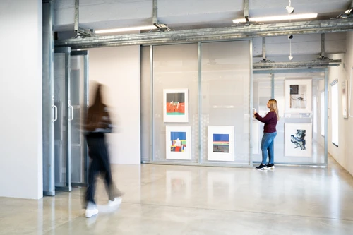 Artothek-Innenraum mit verschiebbaren Gitterpaneelen und gerahmten Werken; zwei Besuchende betrachten die Ausstellung.