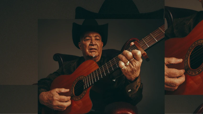 A portrait photograph of musician Elias Ochoa with his instrument, an acoustic guitar, wearing a black suit and a black hat.