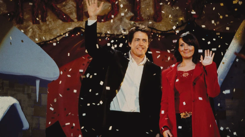 Ein Mann in schwarzem Anzug mit einer Frau in rotem Mantel. Es regnet Papierschnipsel, sie winken