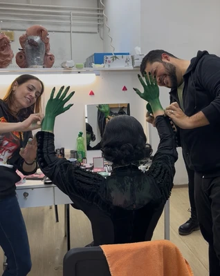 Make-up-Artists Edu Gomes und Flavia Shishov  tragen grüne Farbe auf die Hände und Arme von Laura Panzeri auf, die man von hinten vor einem Spiegel sitzend sieht. Sie streckt die Hände Arme in die Luft, während sie geschminkt werden. Auf dem Tisch liegen verschiedene Kosmetikprodukte, Pinsel und Schwämme, im Hintergrund sind Spiegel und Masken sichtbar.