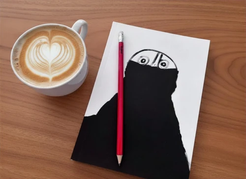 Weiße Kaffeetasse mit Cappuccino auf hellem Holztisch. Daneben liegt ein weißes Blatt Papier mit einer schwarzen, abstrakten Zeichnung, aus der zwei Augen hervortreten. Ein roter Bleistift liegt senkrecht über dem Blatt und teilt die Zeichnung mittig.