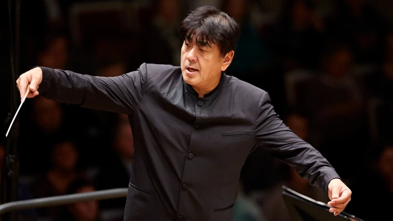 Photo of conductor Yutaka Sado, dressed in a black suit and seen conducting.