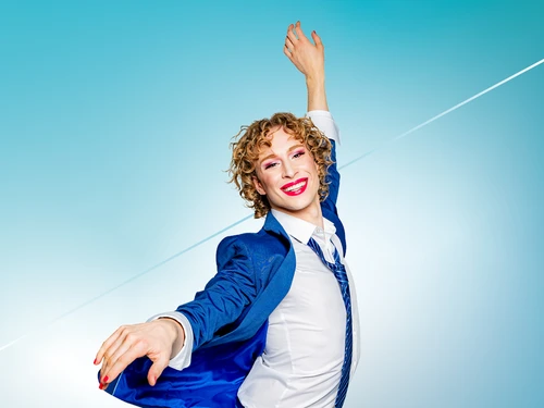 Person in blauem Anzug mit ausdrucksvoller Pose vor blauem Hintergrund; Gesicht unscharf, rote Fingernägel sichtbar.