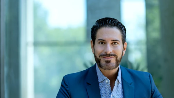 Mann mit dunklen Haaren und dunklem Dreitagebart in dunkelblaumen Anzug mit hellem Hemd vor einem Fenster