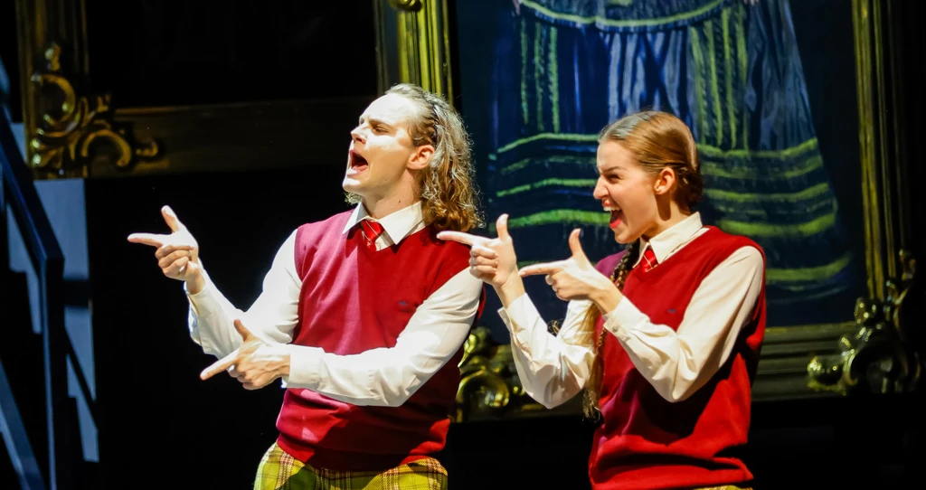 Szenenbild aus dem Musical "Das Gespenst von Canterville". Zwei Personen in roten Pullundern und karierten Hosen stehen auf einer Bühne und zeigen mit den Händen eine Geste wie Fingerpistolen. Im Hintergrund hängt ein großes, gerahmtes Gemälde.