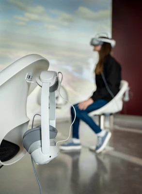A pair of gray VR glasses hangs on an armchair, and in the background, a person wearing VR glasses sits in front of a projection.