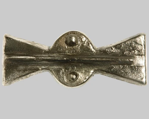 Fibel mit propellerförmiger Kopfplatte. Mittelrippe in Längsrichtung und Buckelverzierung am Mittelteil. Halterung und Einhängevorrichtung für Nadel auf der Rückseite erhalten. Die Nadel fehlt.
