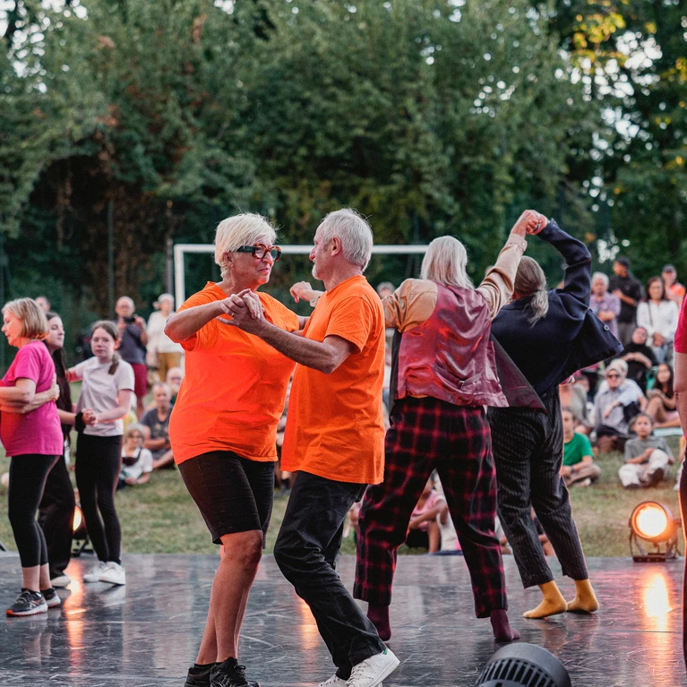 Scenes from a community project about Beauséjour. The picture shows a group of people of all ages on an open-air stage, surrounded by an audience sitting in a meadow. 