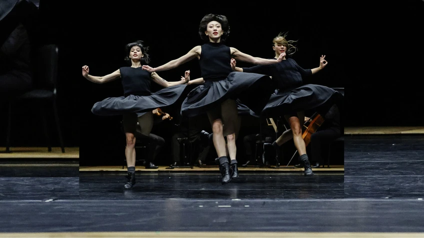 Performancefoto der Choreografie "Bartók" von Anne Teresa De Keersmaeker von 1986 in der sich die Choreografin von Béla
Bartóks "Streichquartett Nr. 4" inspirieren ließ. Zu sehen sind Tänzer:innen des Opera Ballet Vlaanderen in schwarzen Kostümen vor einem schwarzen Bühnenhintergrund in einer dynamischen Gruppenchoreografie, im Hintergrund außerdem ein kleines, musizierendes Kammerorchester.