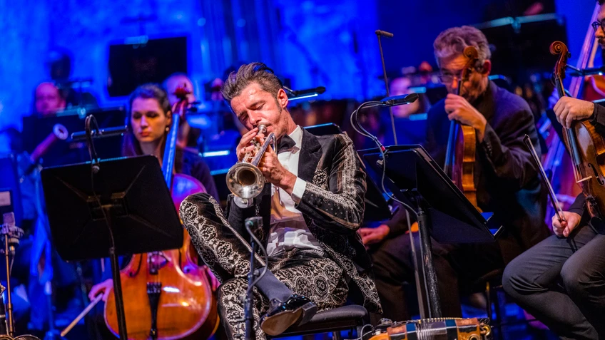 Konzertfoto von Herbert Pixner mit eine Trompete spielend, hinter ihm zu sehen: das Tonkünstler Orchester Niederösterreich auf einer blau beleuchteten Bühne.
