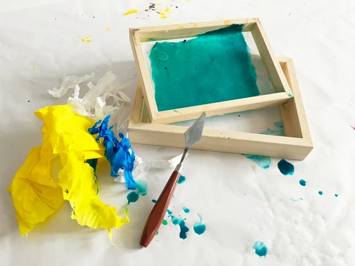 Kleiner Arbeitsbereich mit zwei leeren Holzrahmen, türkis eingefärbtem Papier, gelben und blauen Farbresten sowie einem Spachtel auf weißer Unterlage.