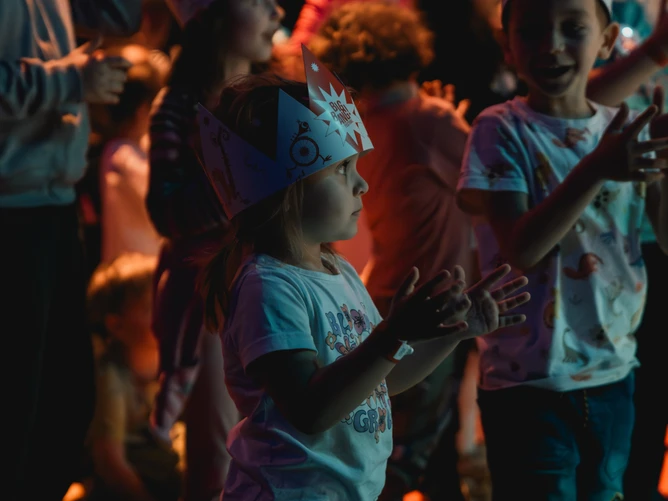 Atmospheric photo from the "BIG BANG Festival – Music Adventure for Young Audiences"; a girl stands in the foreground wearing a "BIG BANG" crown on her head and clapping her hands.