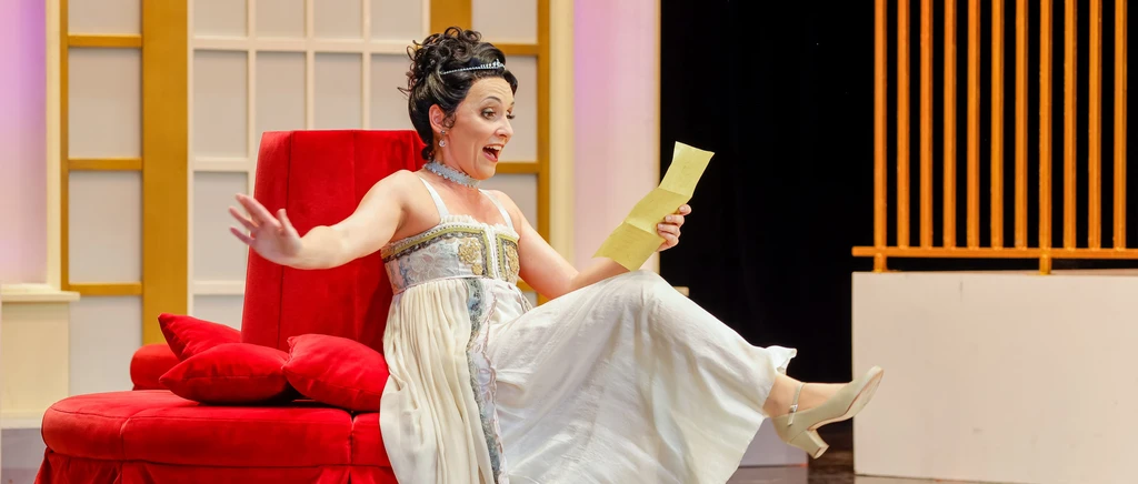 Dame sitzt auf rotem Sessel und ein Bein gehoben, sie trägt ein weißes Kleid und hält einen Zettel in der Hand, ebenfalls streckt sie eine Hand weg
