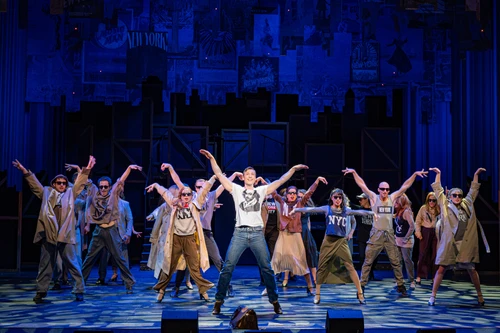 A large ensemble scene on a theatre stage featuring numerous performers spread across the entire width of the stage in several rows. In the foreground, a person in light-coloured clothing stands centrally between the other performers. Many of the performers have both arms stretched diagonally upwards and are striking a synchronised pose. The stage is bathed in a strong blue light and framed by a tall backdrop featuring posters and stage elements arranged one above the other. The patterned stage floor and the group’s uniform posture emphasise the impression of a choreographed dance or musical scene. The central figure forms the visual focal point of the photograph.
